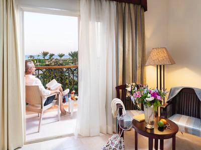 Frau entspannt auf Balkon mit Getränk, Blick auf Meer, gemütliches Hotelzimmer mit Blumen und Lampe.