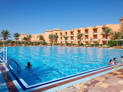Large hotel pool with clear water, surrounded by palm trees and a multi-story building.