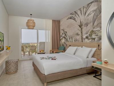 Helles, modernes Hotelzimmer mit Doppelbett, Palmenwandbild und Blick auf einen Balkon.