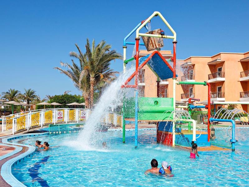 Kinder spielen im Wasserpark mit Rutsche und Wassersprühern neben einem Hotel.