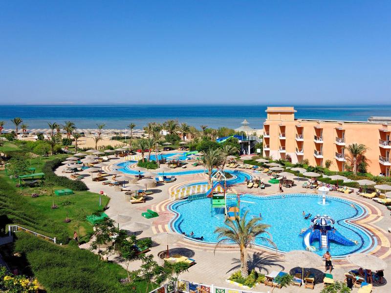 Großer Hotelpool mit Wasserrutsche, Liegestühlen und Blick auf das Meer bei klarem Himmel.
