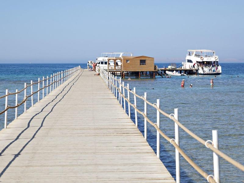 Langer Steg mit Geländer, der zu einem schwimmenden Haus und Boot am Meer führt.