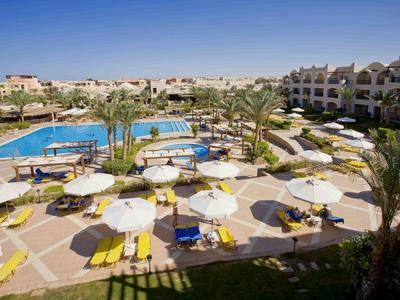 Resort with swimming pools, palm trees, and sun loungers under umbrellas in a sunny courtyard.