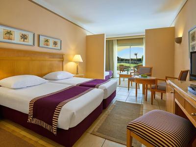 Hotel room with two beds, purple bed runners, desk, TV, and balcony with outdoor view.