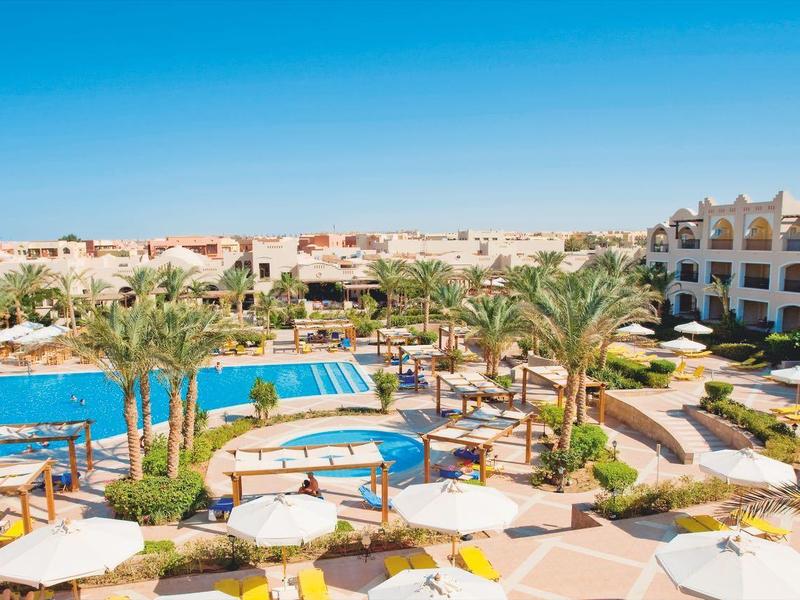 Resort with two blue pools, white umbrellas, palm trees, and beige buildings under a clear sky.