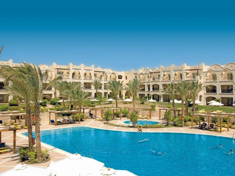 Large hotel with swimming pool, palm trees, and lounge chairs under a blue sky.