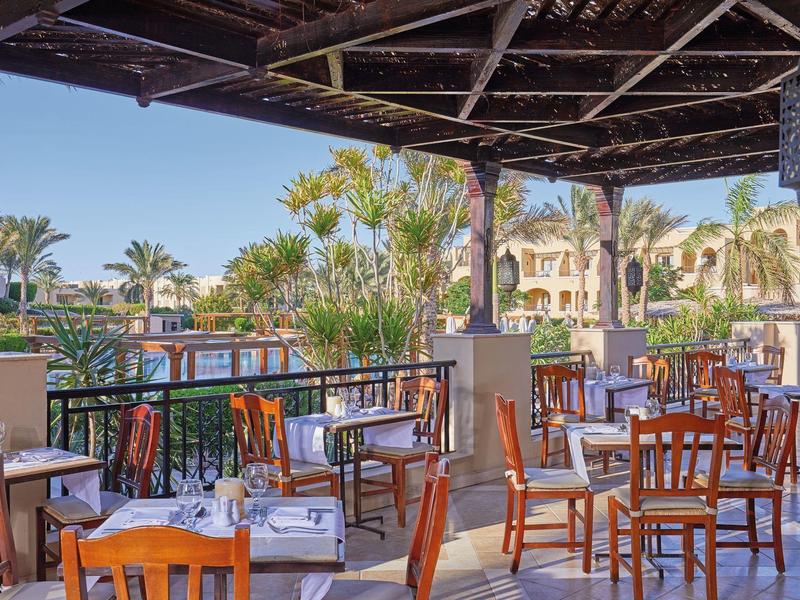 Outdoor dining area with wooden tables and chairs overlooking a pool and tropical plants.