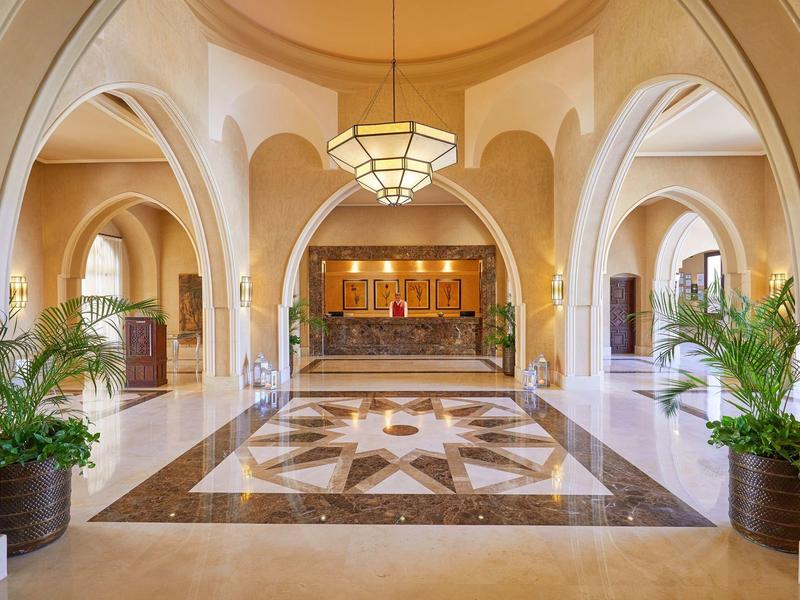 Spacious hotel lobby with arched doorways, marble floors, large chandelier, and potted plants.
