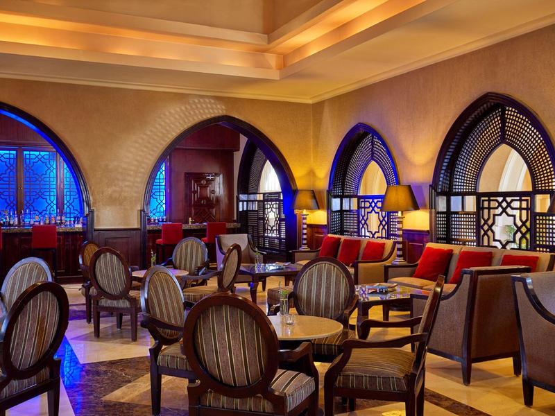 Cozy hotel lounge with elegant wooden chairs, tables, arched windows, and warm lighting.