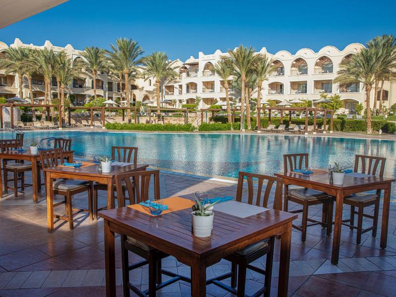 Hotel terrace with tables and chairs by the pool, surrounded by palm trees and a white building.