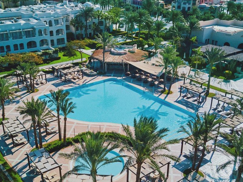 View of a large hotel pool with sun loungers and palm trees in a tropical setting.