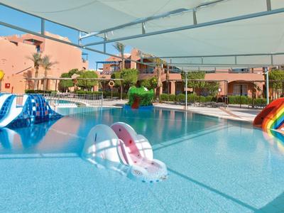 Großer Pool mit bunten Wasserrutschen und Sonnensegel, umgeben von rosa Gebäuden.