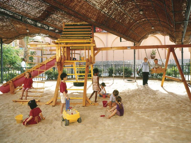 Kinder spielen im Sandkasten unter einem schattigen, aus Holz und Stroh gebauten Spielplatz.