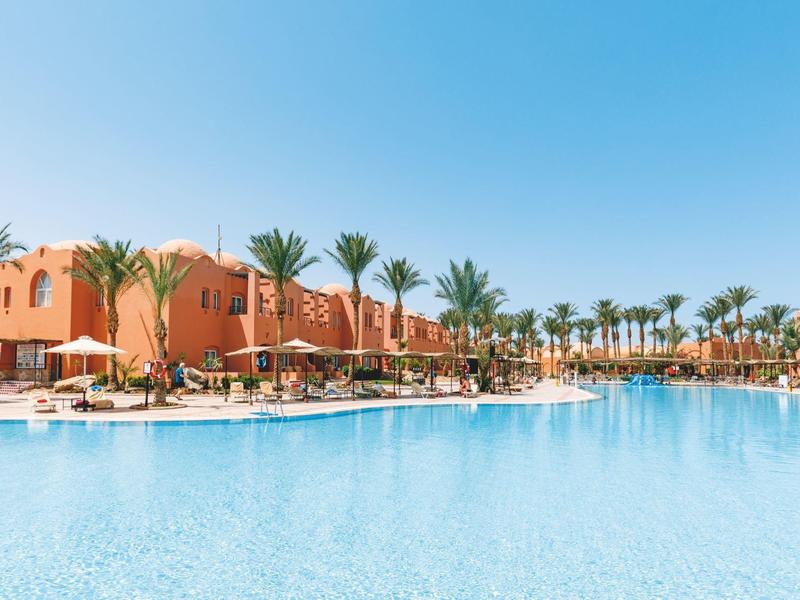 Großer blauer Pool vor orangen Häusern, umgeben von Palmen und Sonnenschirmen bei klarem Himmel.