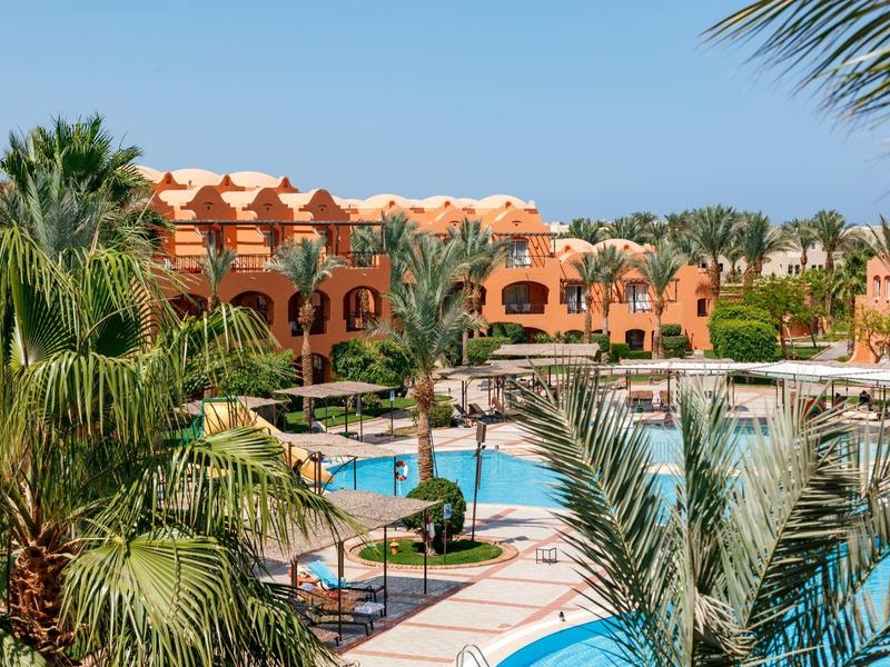 Hotelanlage mit orangefarbenen Gebäuden, grünen Palmen und mehreren blauen Pools bei klarem Himmel.