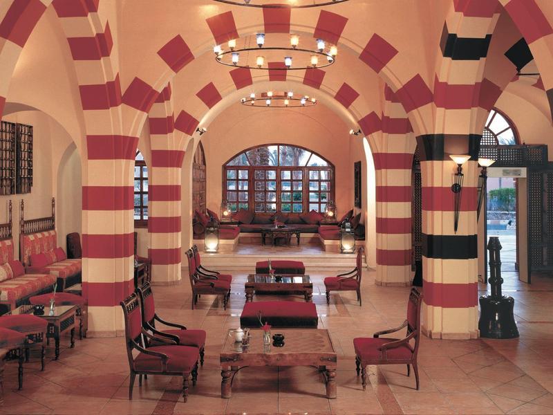 Innenraum mit roten und weißen Bogen, Holzmöbeln und rechteckigen Tischen in warmem Licht.