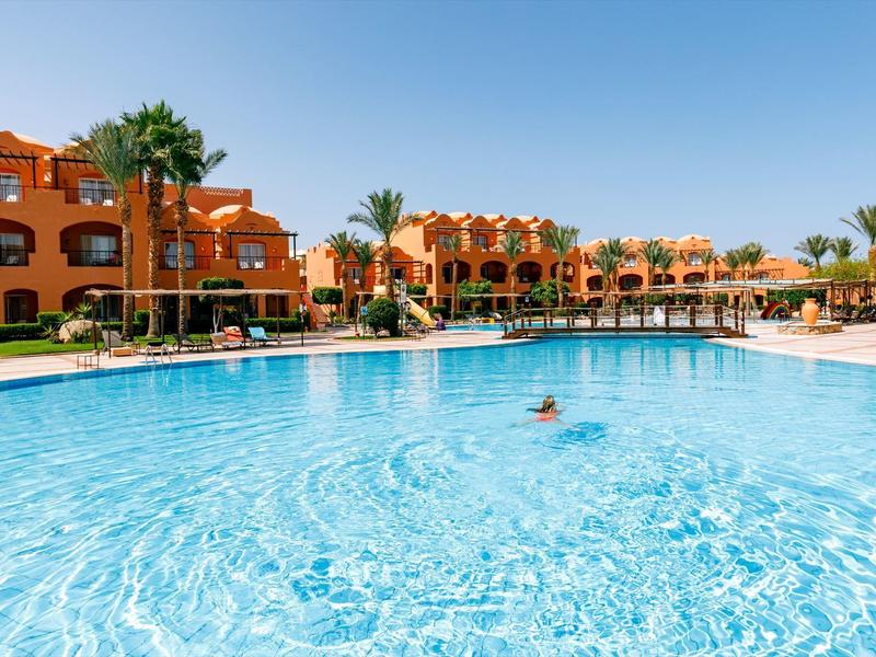 Großer blauer Pool vor orangefarbenen Gebäuden mit Palmen und klarem Himmel.