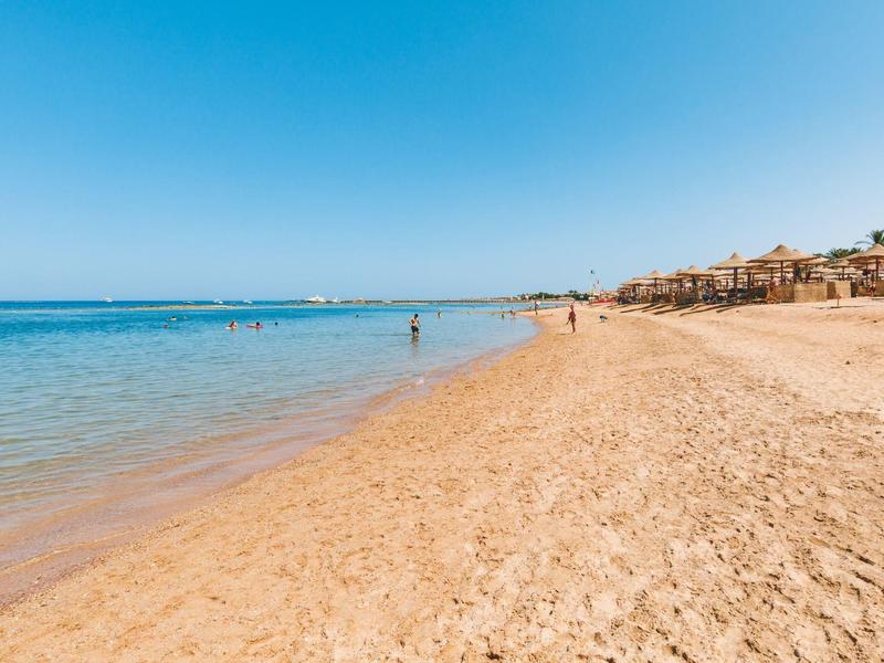 Weiter Sandstrand mit klarem Wasser, blauer Himmel und vereinzelten Menschen am Strand und im Wasser.
