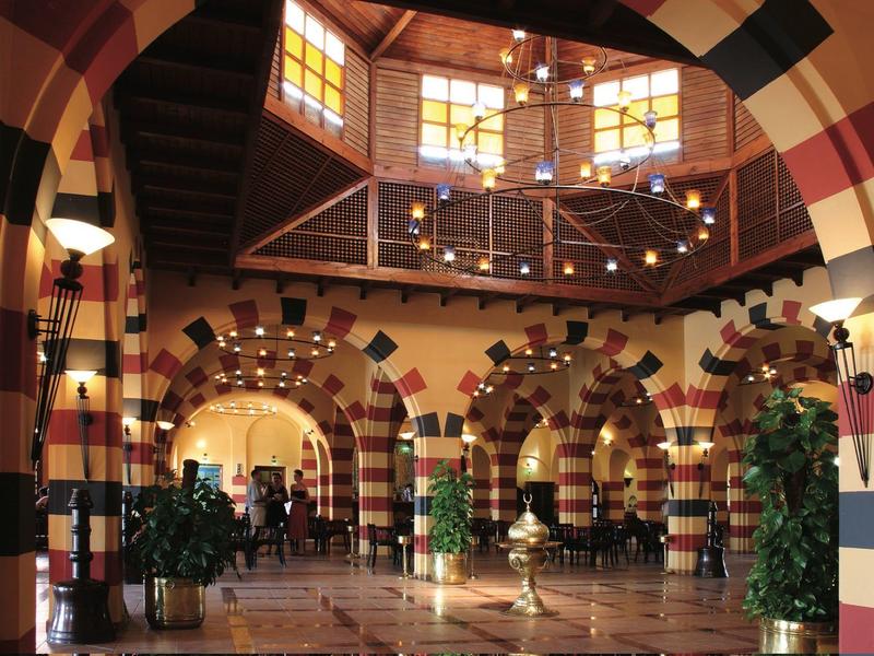 Große Halle mit gestreiften Steinbögen, Holzbalkon, Glasfenstern und mehreren Pflanzen.