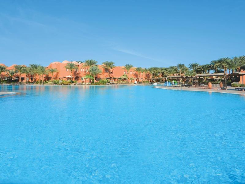 Großer Pool vor roten Gebäuden und klarem blauem Himmel in einem Resort.
