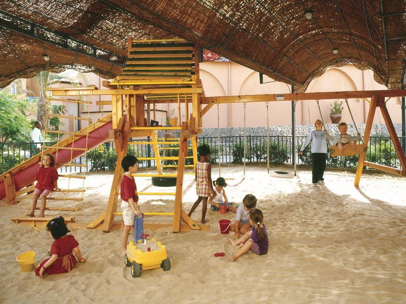 Kinder spielen auf einem schattigen Sandspielplatz mit Rutschen und Schaukeln.