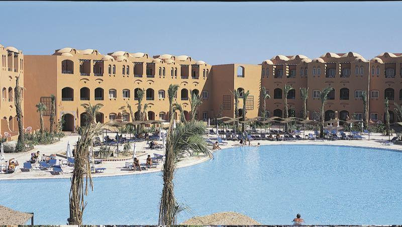 Großer blauer Pool umgeben von Palmen und mehrstöckigen Sandstein-Gebäuden unter klarem Himmel.