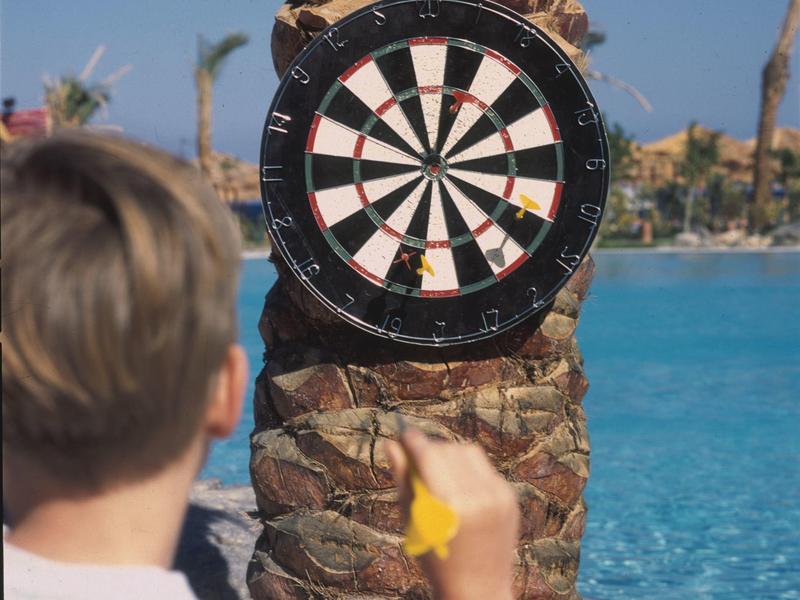 Ein Mann wirft Darts auf eine Dartscheibe, die an einem Baumstamm neben einem Pool befestigt ist.