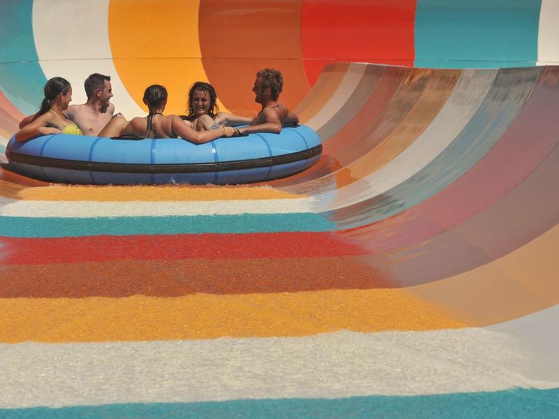 Fünf Personen in einem runden Schlauchboot auf buntem Wasserrutschen-Bogen mit kräftigen Farben.