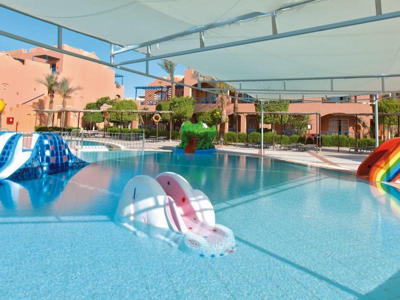 Großer Pool mit bunten Wasserrutschen und Sonnensegel, umgeben von rosa Gebäuden.