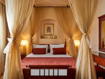 Gemütliches Schlafzimmer mit Himmelbett, roten Akzenten, warmem Licht und Gardinen aus hellem Stoff.