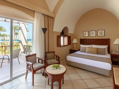 Gemütliches Hotelzimmer mit Doppelbett, Sitzbereich, Balkon und Blick auf Palmen und Pool.