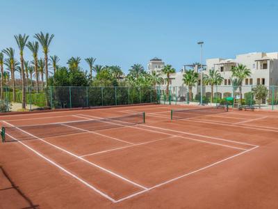 Roter Sand-Tennisplatz mit Netz, umgeben von Palmen und weißen Gebäuden bei klarem Himmel.