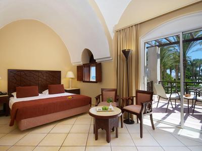Hell eingerichtetes Hotelzimmer mit großem Bett, Sitzbereich und Balkon mit Blick auf Palmen.