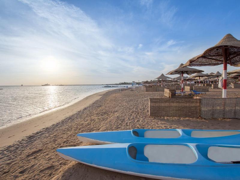 Blauer Katamaran auf Sandstrand mit Sonnenschirmen und ruhigem Meer bei Sonnenuntergang.