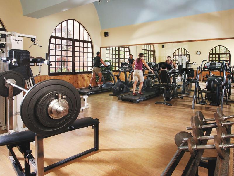 Ein heller Fitnessraum mit Holzfußboden, Trainingsgeräten und mehreren Personen beim Sport.