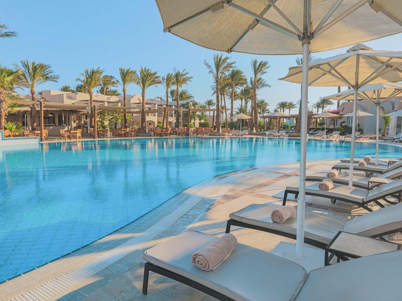 Großer Pool mit Liegestühlen und Sonnenschirmen, umgeben von Palmen und Hotelgebäuden.