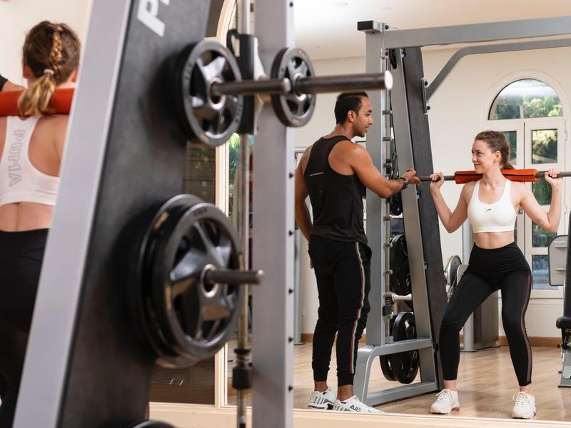 Man coaches woman lifting weights in gym with mirrors and equipment around.