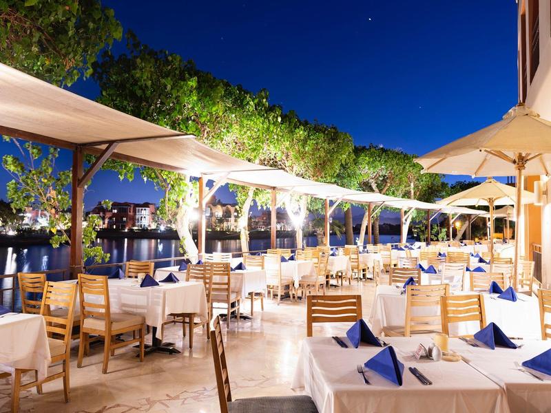 Outdoor restaurant with tables, chairs, umbrellas and ambient evening lights near water.