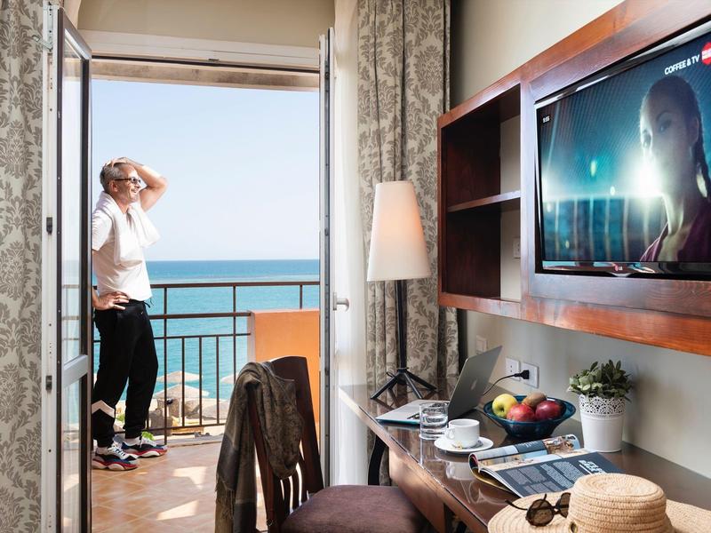Hotel room with balcony overlooking the sea, man enjoying the view and TV on the wall.