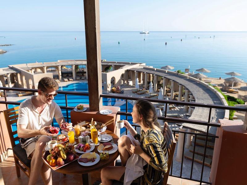 Couple enjoys breakfast on balcony overlooking beach and ocean with clear blue sky.