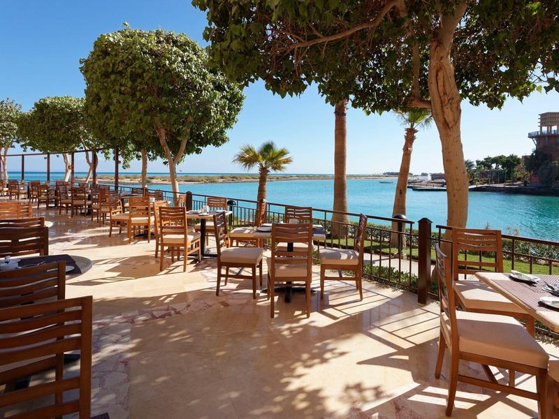 Outdoor seating area with wooden tables and chairs near the sea under leafy trees.