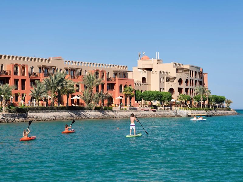 People paddleboarding near a coastal hotel with clear blue water and sky.