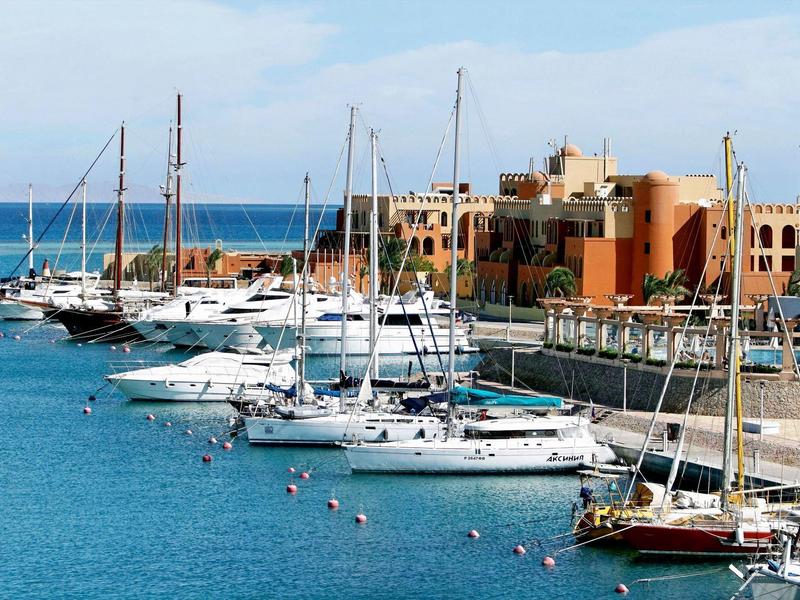 Yachts docked at a marina with buildings and clear blue sea in the background.