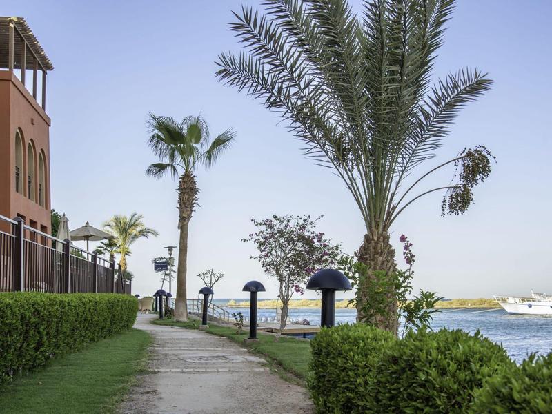 Pathway along waterfront with palm trees, shrubs, and buildings under clear sky.