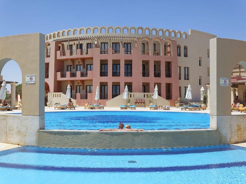 Outdoor swimming pool with loungers and a luxury hotel building under clear blue sky.