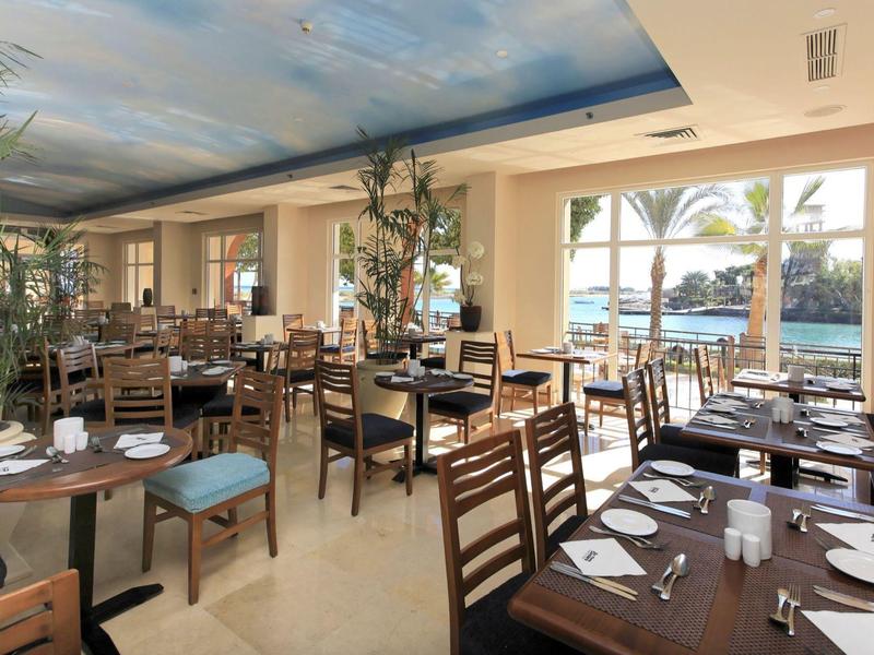 Spacious restaurant with wooden chairs and tables, large windows showing pool and palm trees outside.