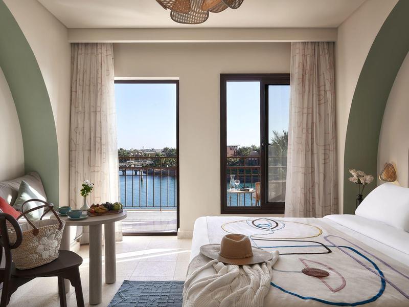 Bright hotel room with double bed, wooden chair, and balcony overlooking the sea.