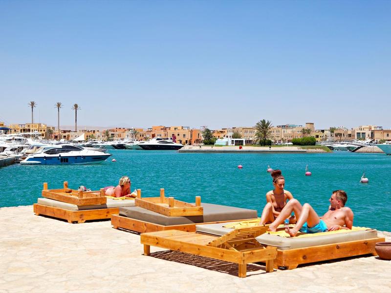 People relaxing on wooden sunbeds by the clear blue sea and boats at a sunny beach.