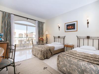 Zimmer mit zwei Einzelbetten, hellen Wänden, Schreibtisch, Stühlen und Balkon mit Glasfenstern.