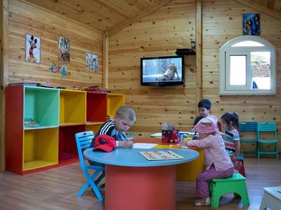 Children play at a colorful round table in a wooden playroom with shelves.